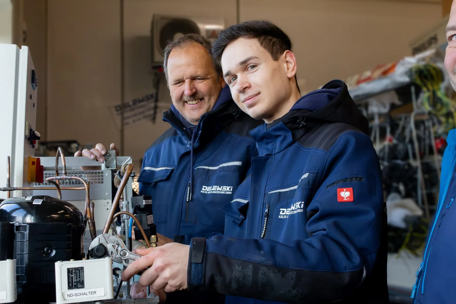 Ein älterer und ein jüngerer Kollege lächeln an einer Kälteanlage in der Werkstatt