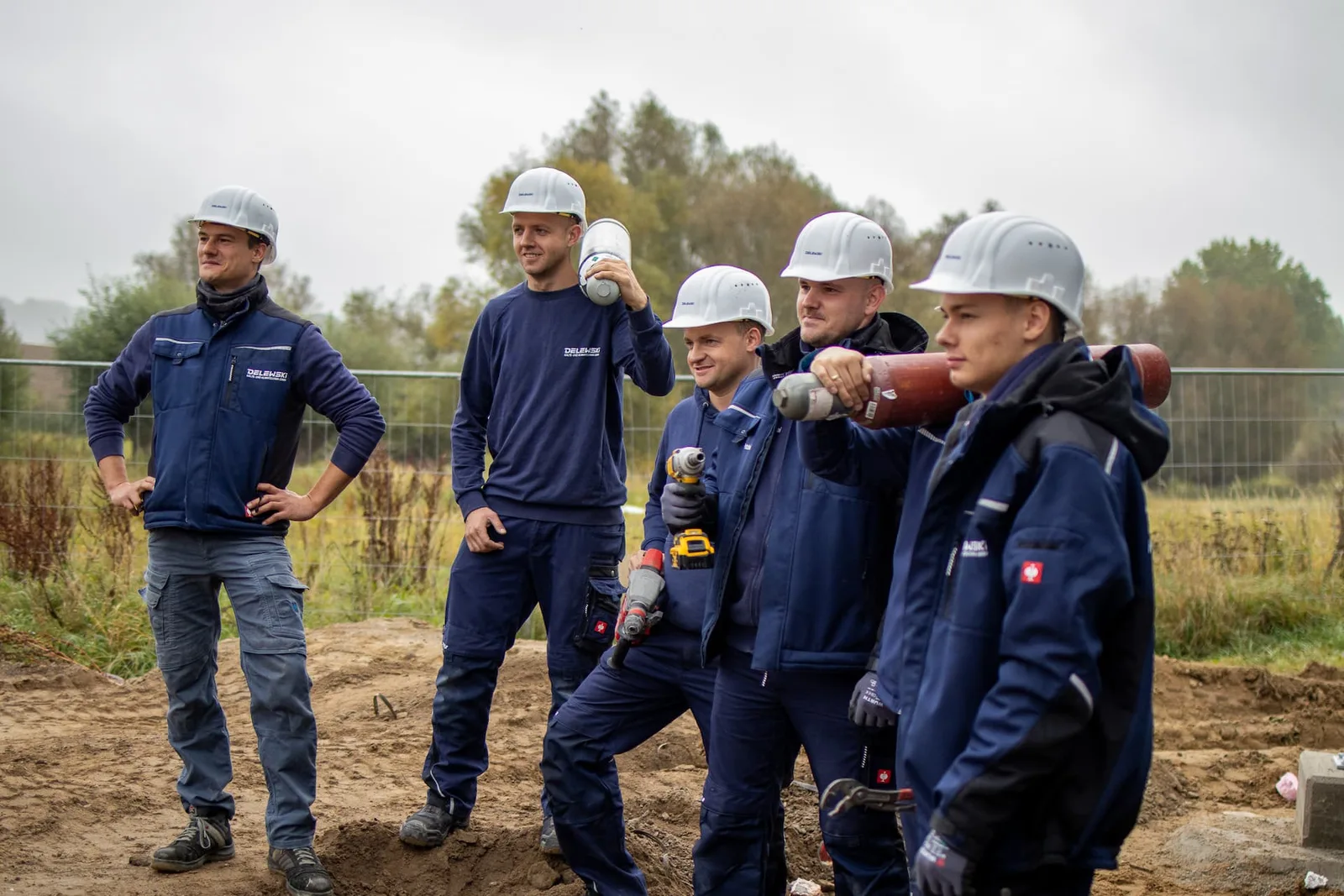 Fünf Monteure mit Helmen auf der Baustelle, zwei tragen Rohre auf der Schulter