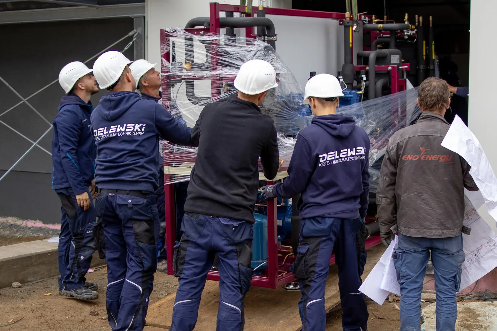 Monteure in Delewski-Jacken schieben eine in Folie verpackte Anlage über die Baustelle