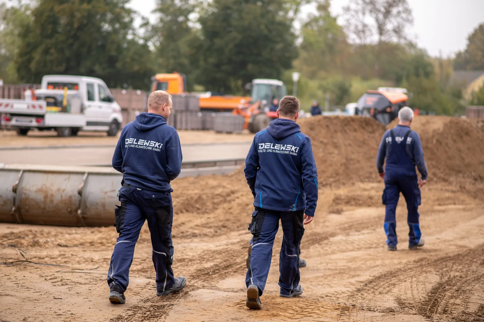 Drei Monteure in Delewski-Jacken gehen über den sandigen Baustellenboden