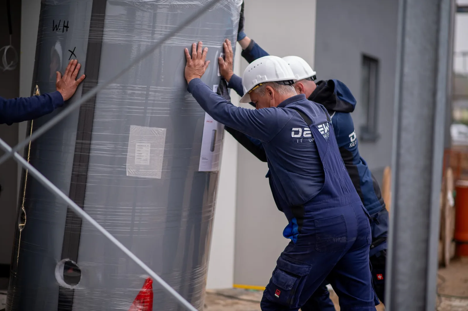 Monteure mit Helm richten einen großen grauen Behälter an der Gebäudewand aus