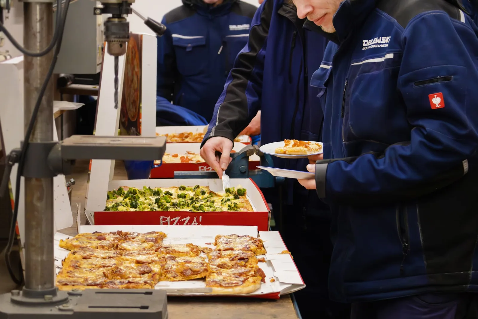 Kollegen in Arbeitsjacken nehmen sich Pizza von einem Tisch in der Werkstatt